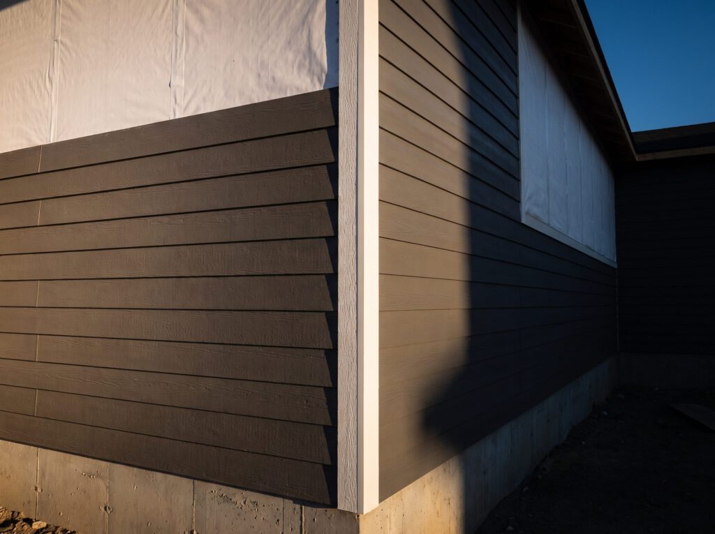 Charcoal fiber cement lap siding installed in even courses up to a white corner board on a new home, with white house wrap still showing above the top course, lit by low evening sun.